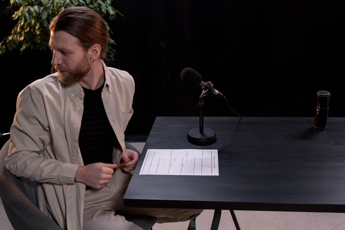 A Man in Brown Long Sleeves Sitting on a Chair Near Microphone on Wooden Table